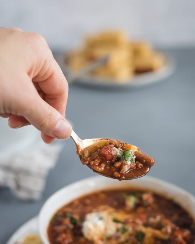 hand holding up spoonful of chili with bowl and plate of cornbread in background