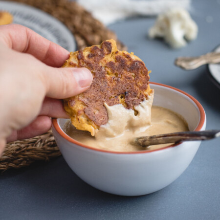 A hand dips a browned fritter into a bowl of creamy sauce with a spoon inside.