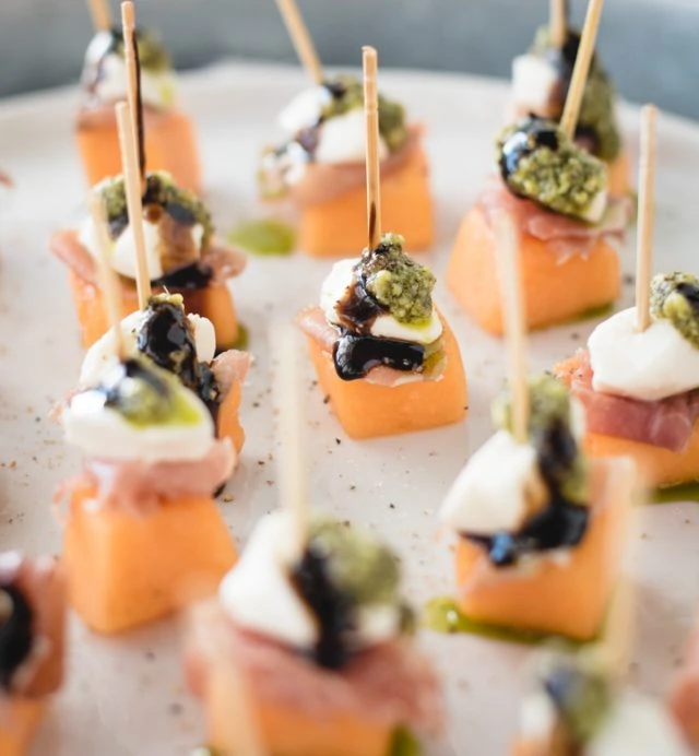 close up picture of a tray of melon, mozzarella, prosciutto skewers topped with pesto and balsamic glaze