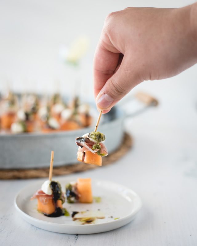 hand grabbing toothpick prosciutto melon skewer off a plate with more in background 