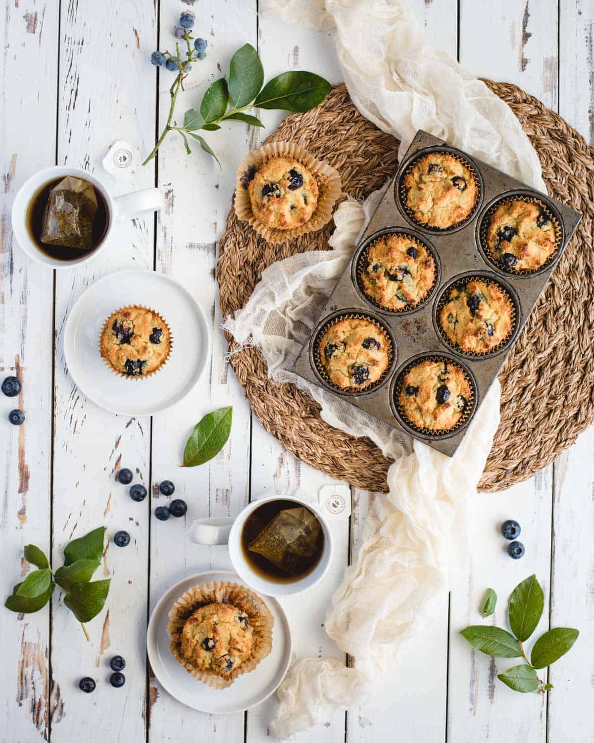 Table scape with two plated blueberry muffins, 6 more muffins in the baking pan, and two mugs of hot tea.