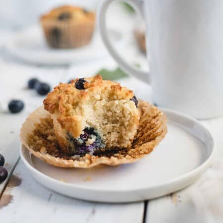 A partially eaten blueberry muffin sits on a white plate next to a mug.