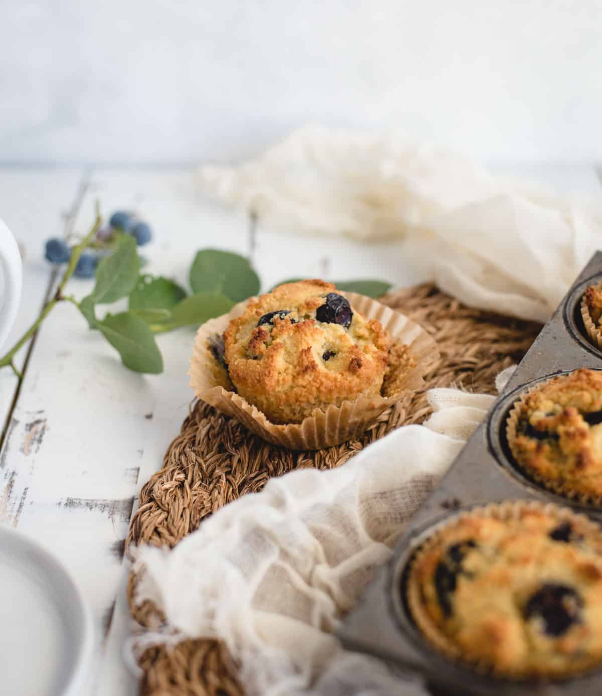 Blueberry muffin in a muffin wrapper sitting on a table right outside a pan of blueberry muffins