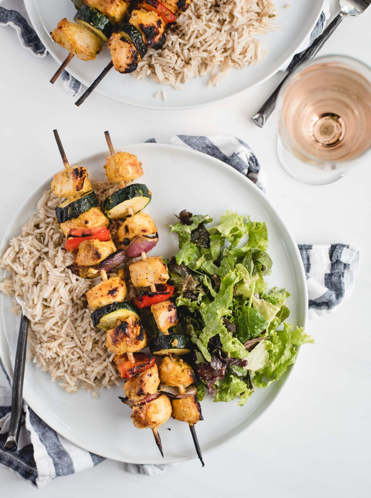 close up overhead picture of two plates of chicken kabobs, salad, and rice with white wine on a white table