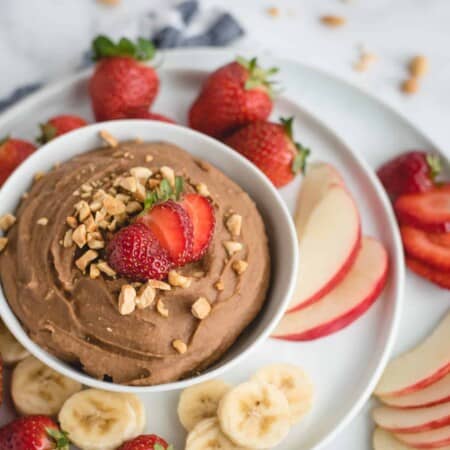 A bowl of chocolate dip topped with chopped nuts and strawberries, surrounded by sliced apples, bananas, and whole strawberries on a white plate.