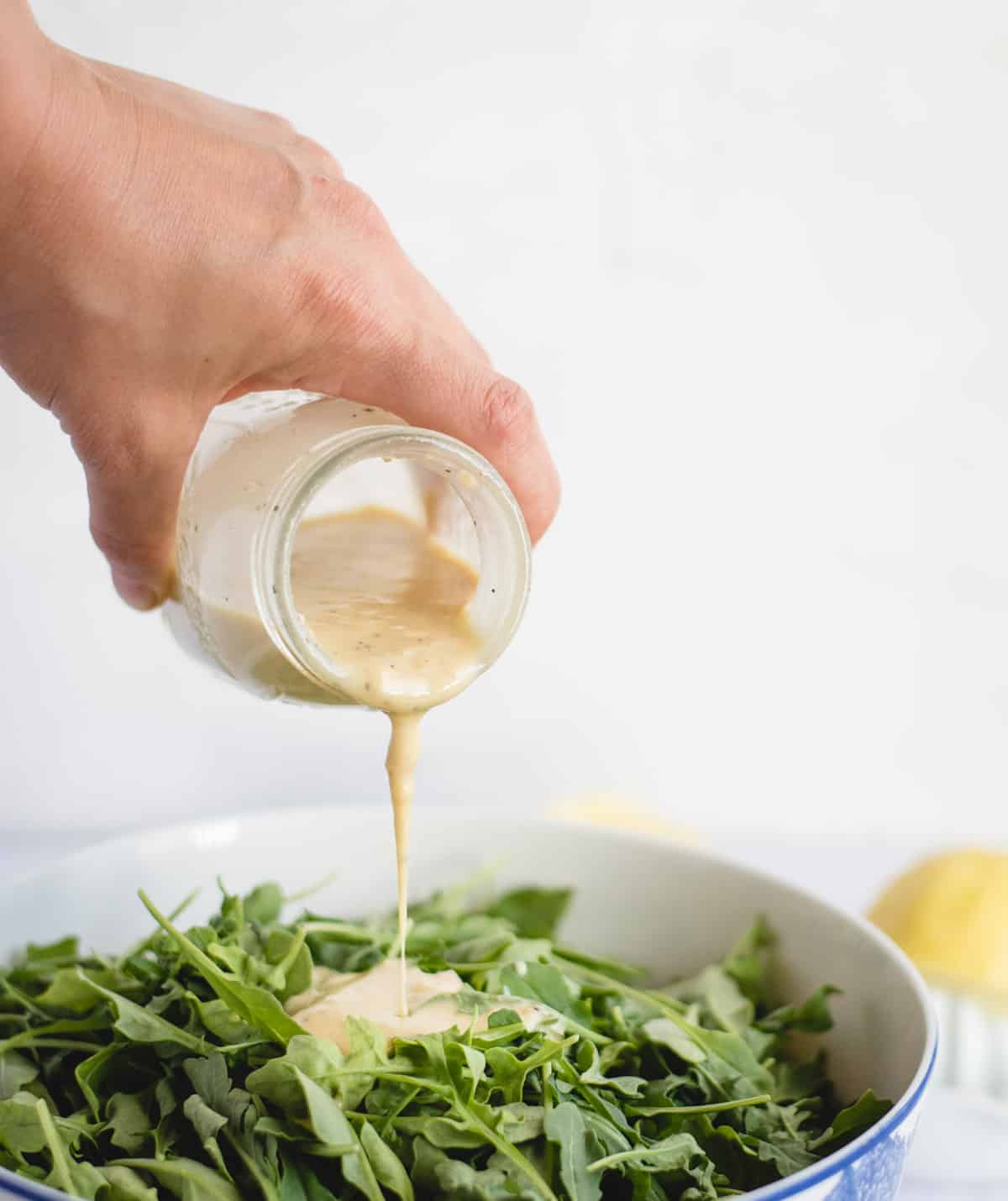 hand pouring lemon tahini salad dressing over a green salad against a white background