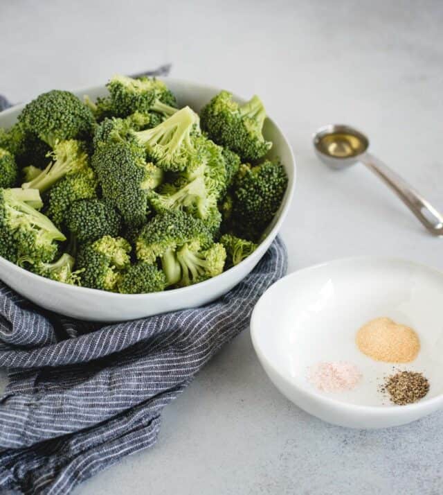 bowl of broccoli, salt, pepper, and garlic powder and a spoon full of oil on a light gray background 