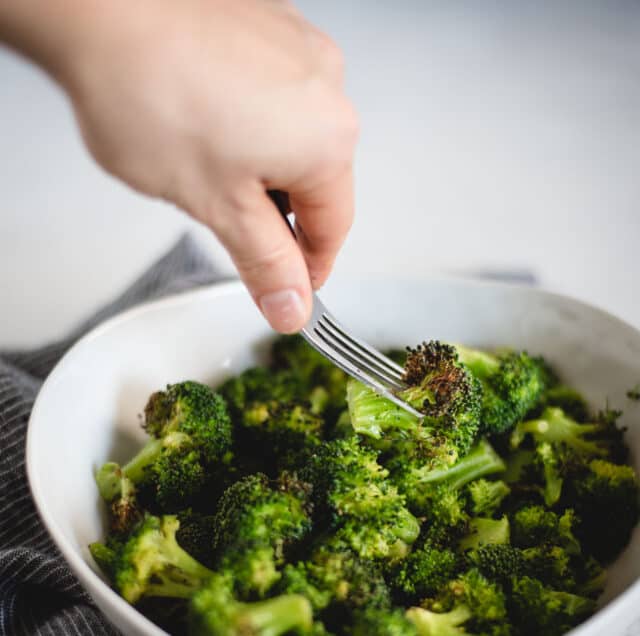 hand reaching fork into a white bowl of cooked broccoli and stabbing a piece