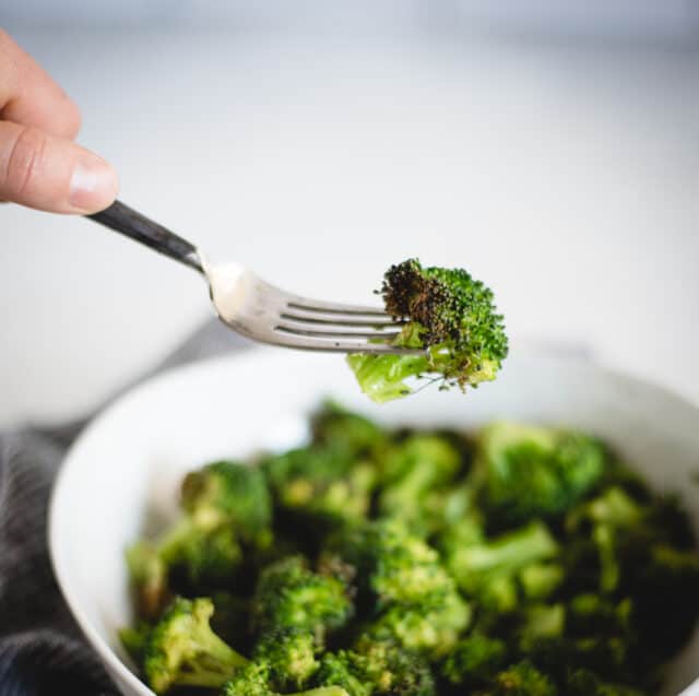 Hand holding a fork with a piece of broccoli on it and a bowl of broccoli in background 