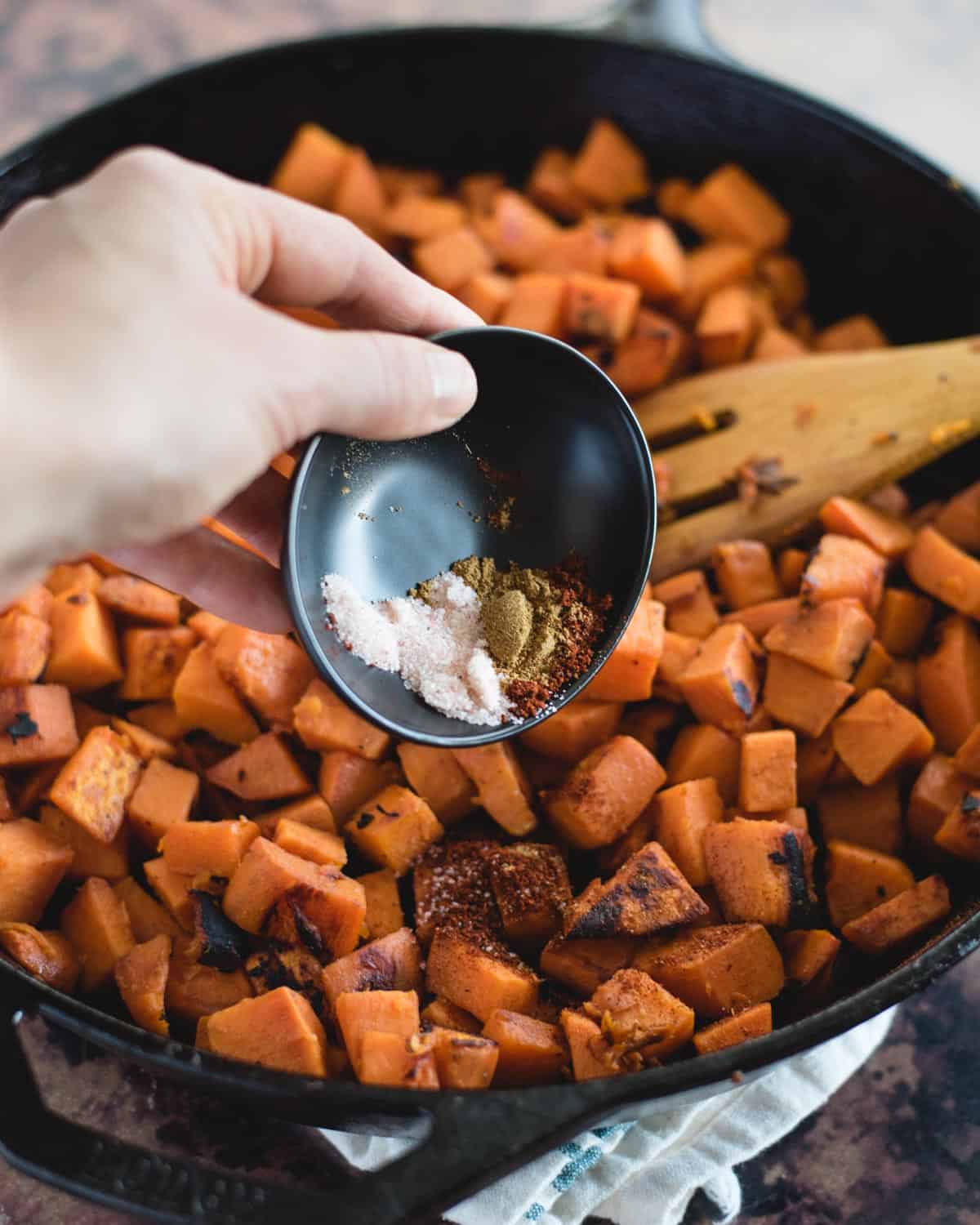 hand pouring seasonings onto a skillet of cooked sweet potato cubes
