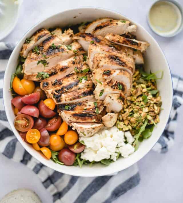 overhead picture of green salad with sliced, grilled chicken, pistachios, tomatoes, and feta on a white background