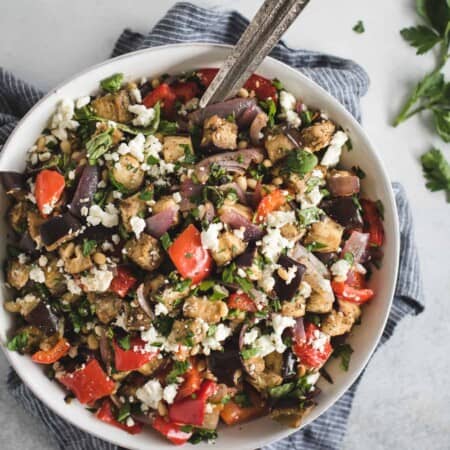 A bowl of Mediterranean salad with grilled vegetables, feta cheese, pine nuts, and fresh herbs on a striped cloth.