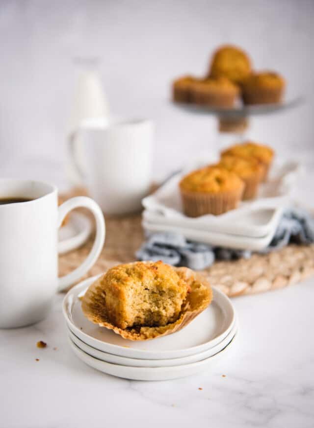 picture of coconut muffin on a white plate with bite taken out and serving plate with muffins in the background