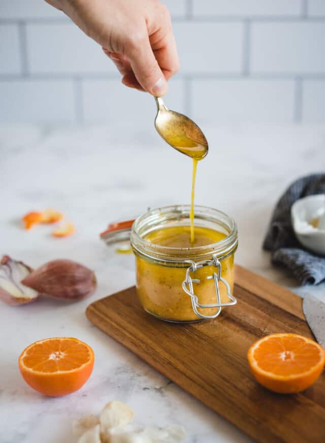Hand holding a spoon pouring orange salad dressing back into its jar on a white background
