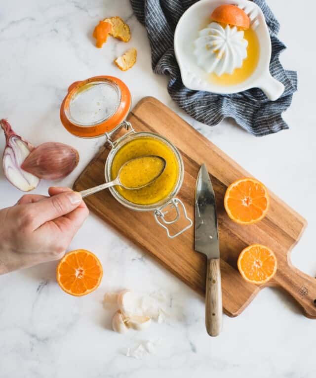 Hand holding spoon of dressing over jar of orange vinaigrette with ingredients all around it 