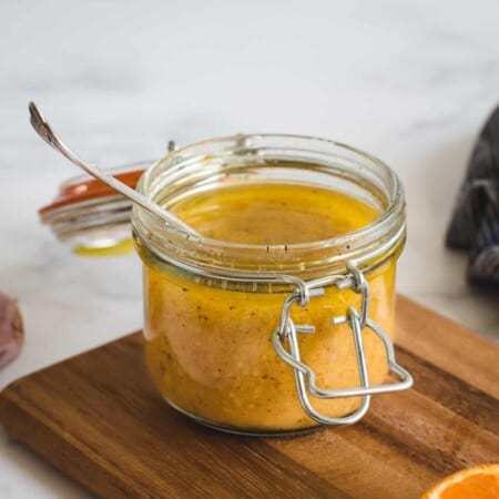 A glass jar filled with mustard dressing sits on a wooden board. A spoon is placed inside the jar.