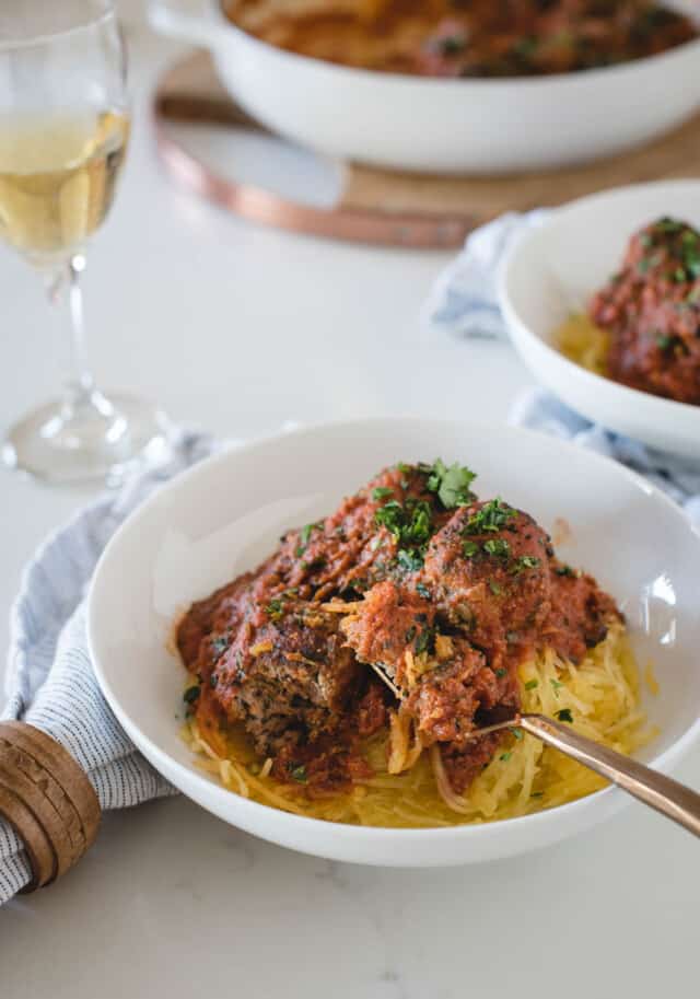 bowl of meatballs in sauce on top of spaghetti squash on a white background with a glass of wine