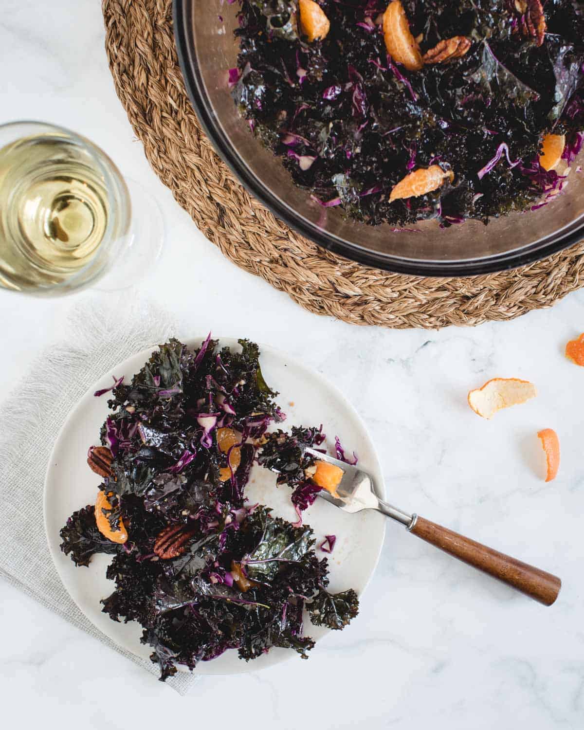 overhead angle of orange kale salad on a plate next to a glass of wine with a larger serving bowl of salad next to it