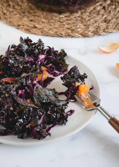 A plate of dark leafy greens, likely kale, mixed with purple cabbage and orange segments, on a white marble surface. A fork rests on the plate, and orange peels are scattered nearby.
