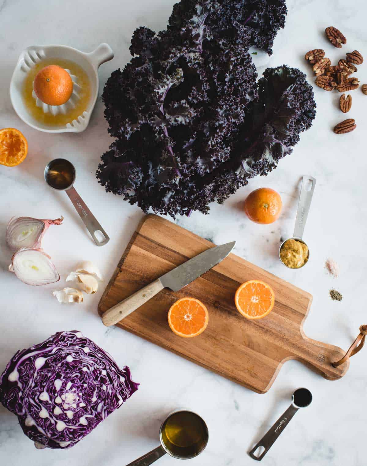 ingredients for kale salad laid out on a marble backdrop including kale, red cabbage, shallot, mandarin oranges, mustard, pecans, oil, and other seasonings
