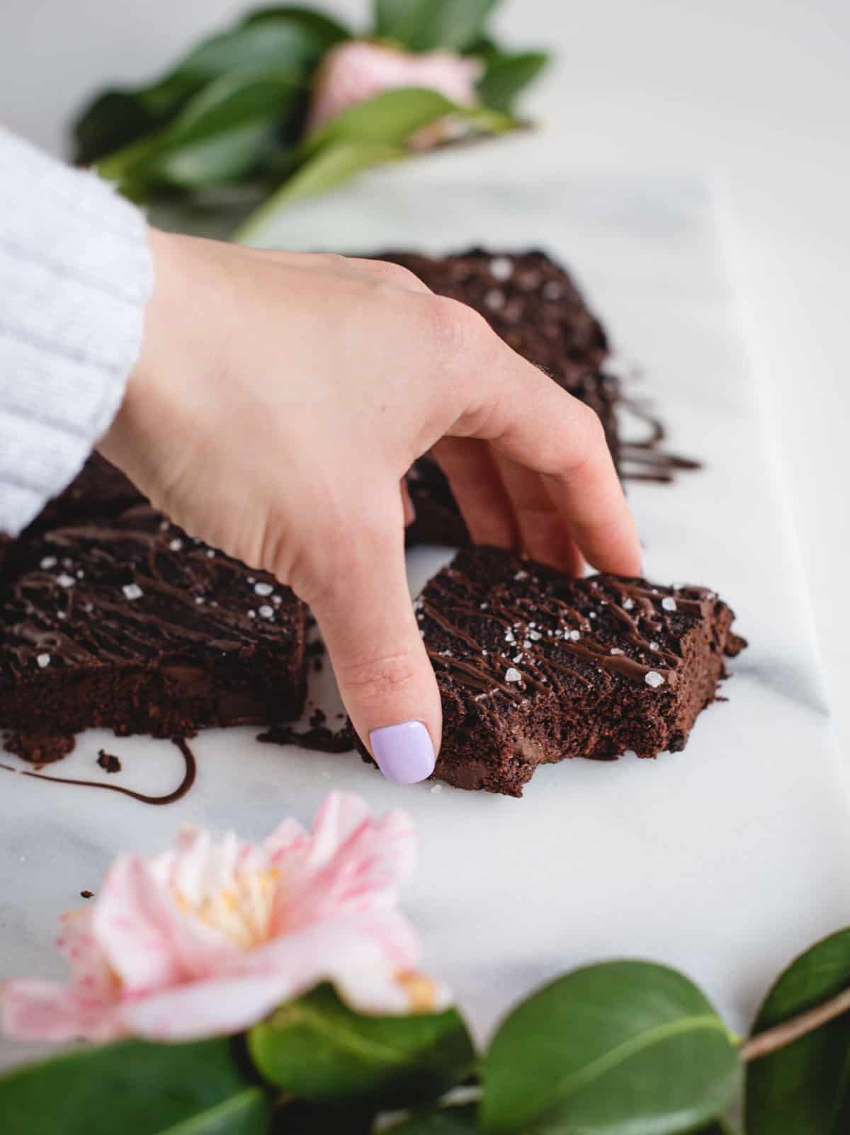 Hand reaching for a healthy banana brownie on a white marble background with pink flowers on edge