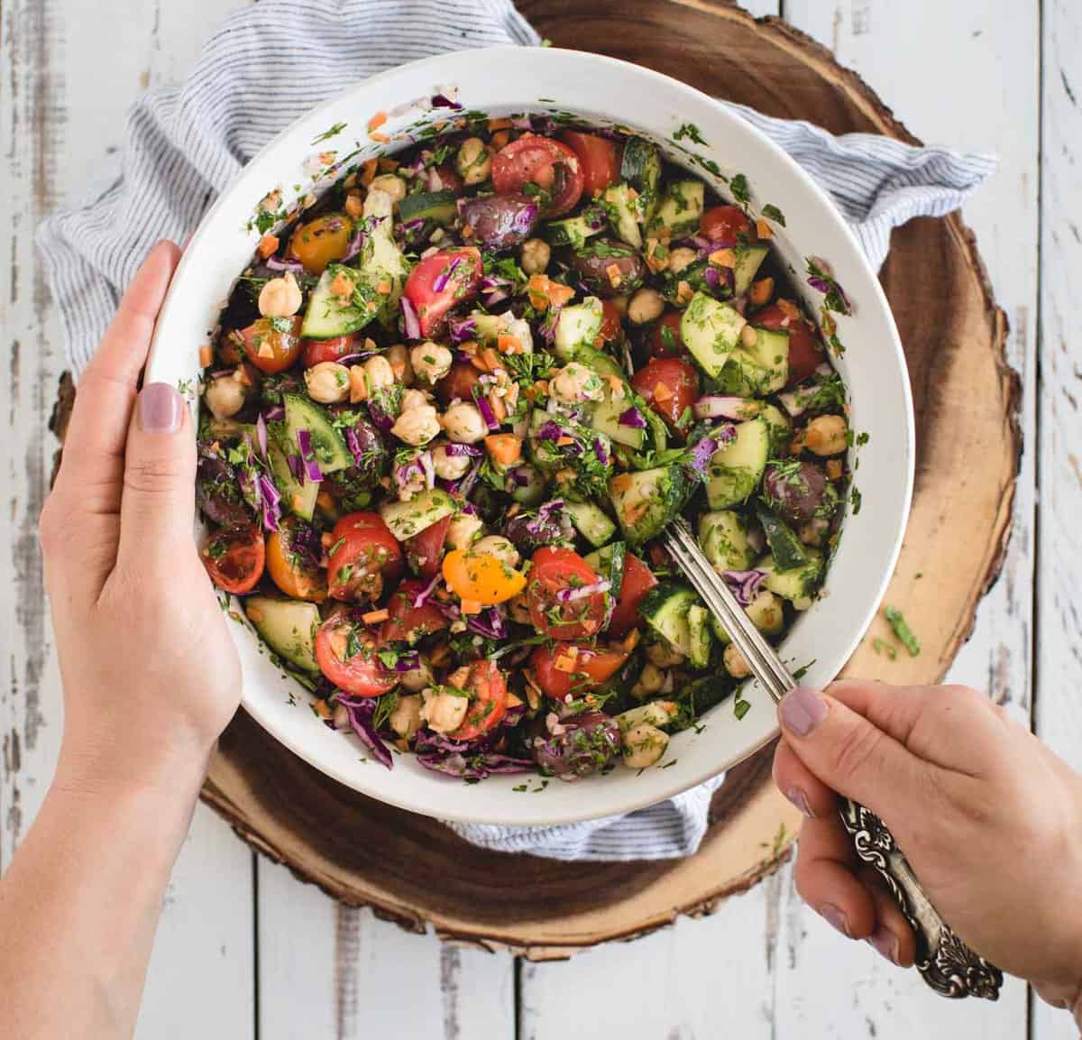 Hand holding bowl and other hand stirring vegan chickpea salad with bright colorful ingredients