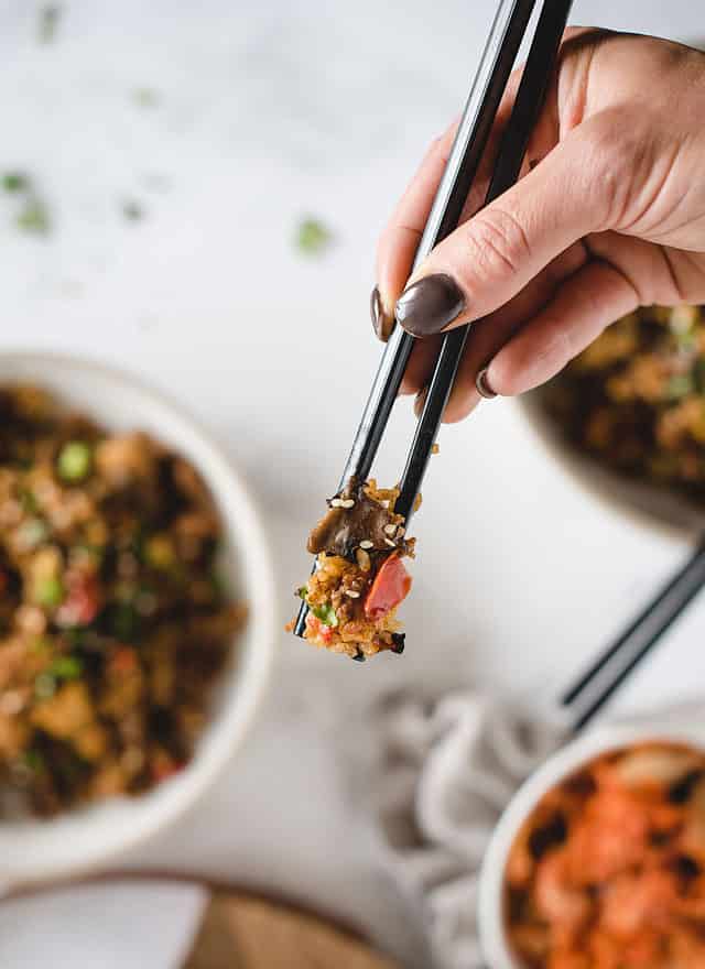 chopsticks holding up bite of veggies and rice with sesame seeds and food in background