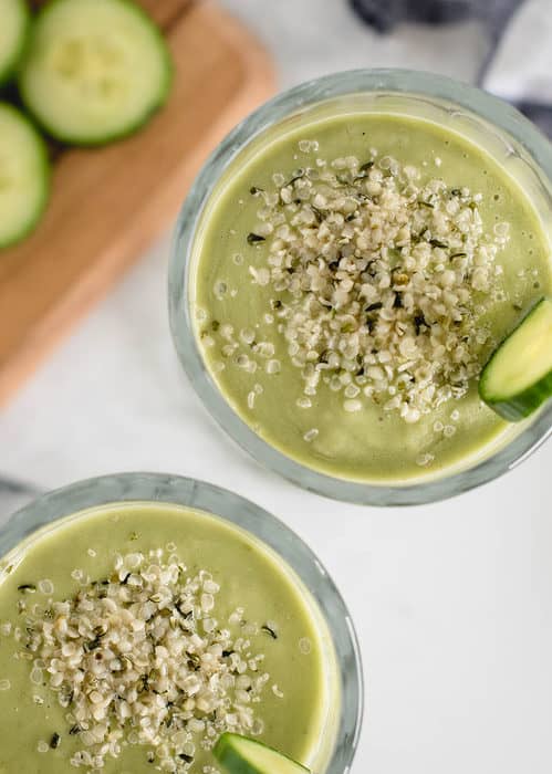 vertical close up overhead of green smoothies with hemp hearts sprinkled on top and cucumber slice on each glass on white background