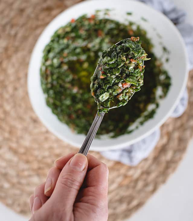 Hand lifting spoon of chimichurri up with bowl of more in the background
