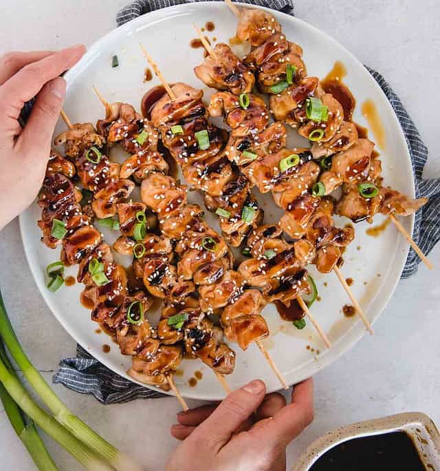 Hands holding a plate full of teriyaki chicken kabobs with green onions