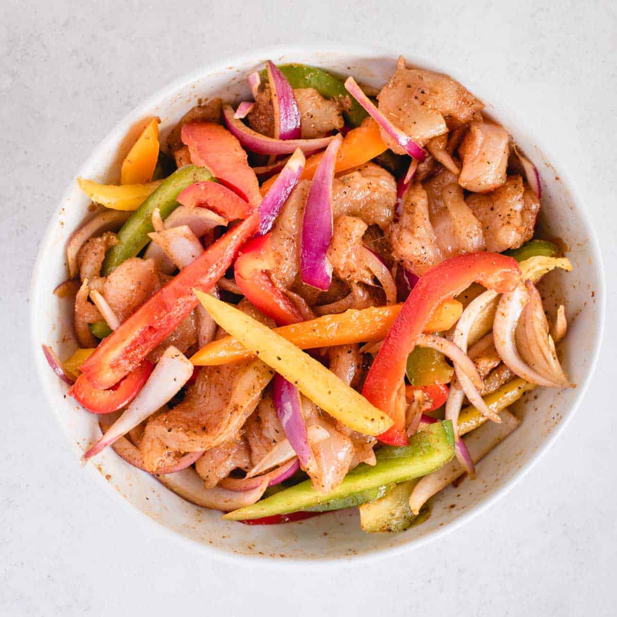cut up raw peppers, red onion and chicken tossed in seasoning in a bowl