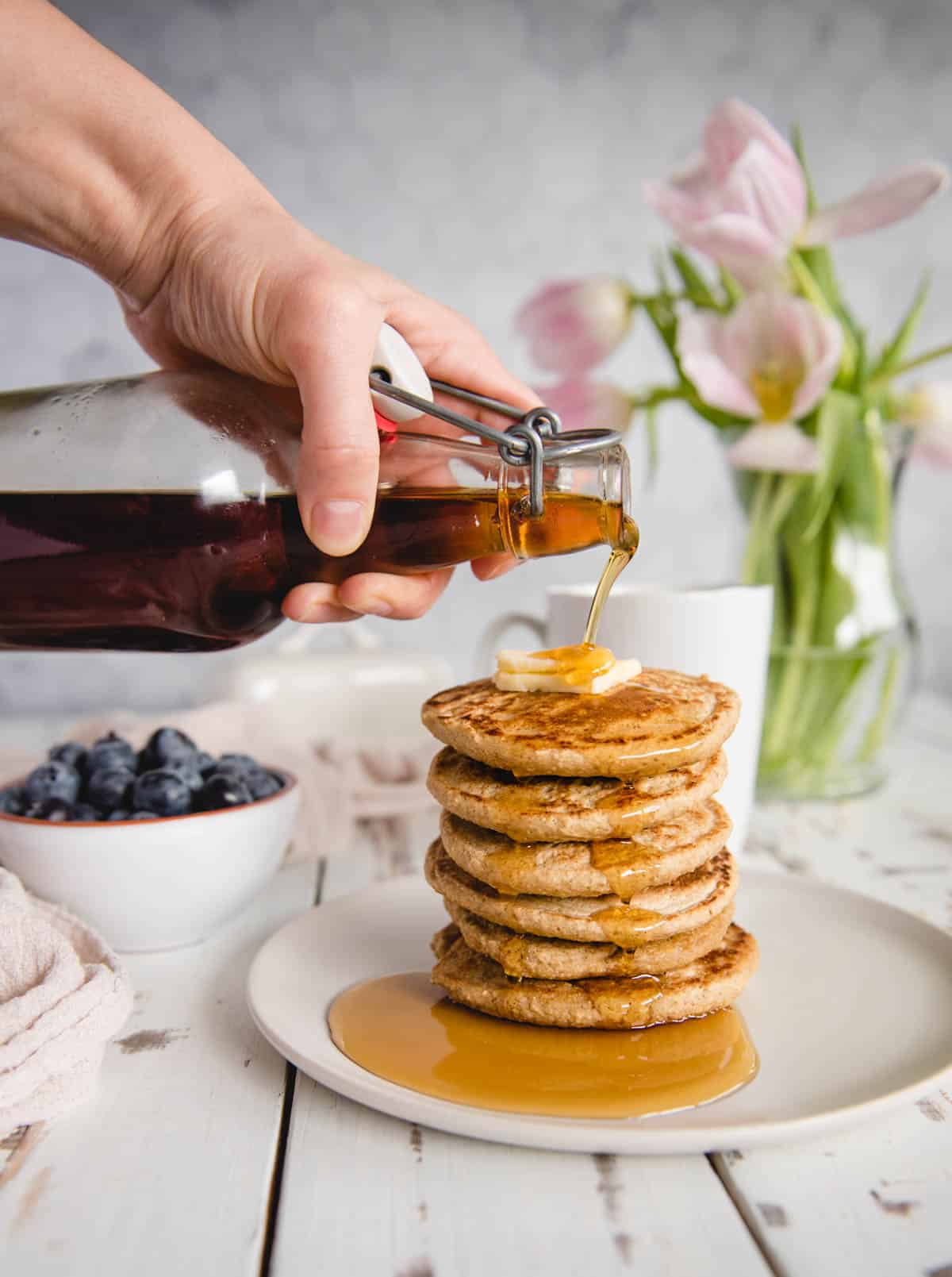 hand pouring syrup from a glass bottle onto a plated stack of gluten free pancakes.