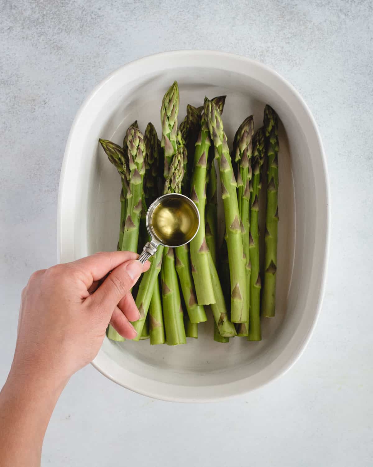 hand holding measuring spoon full of oil over asparagus in a bowl