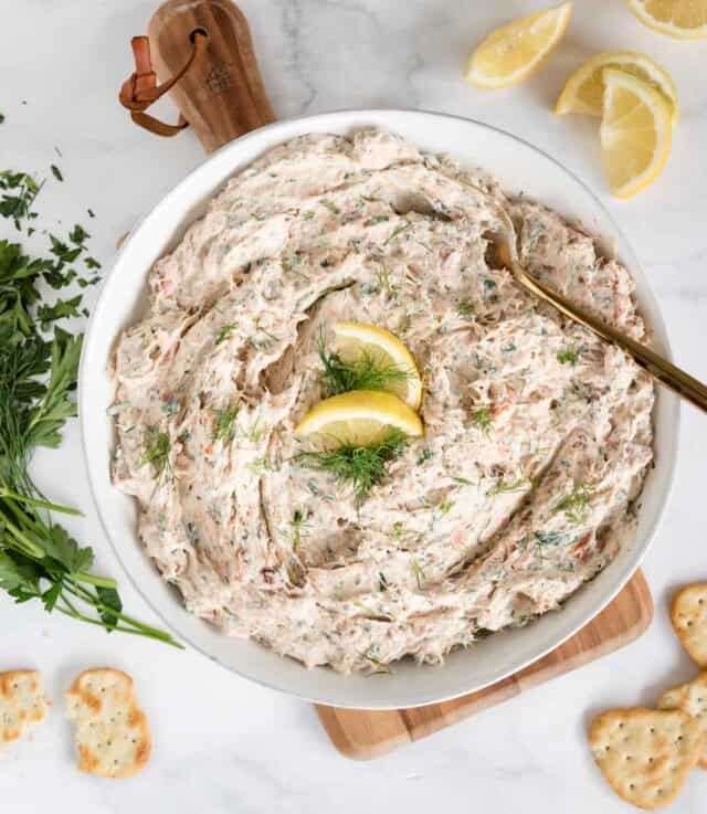 overhead view of white bowl of smoked salmon cream cheese based dip with crackers, lemon and herbs next to it