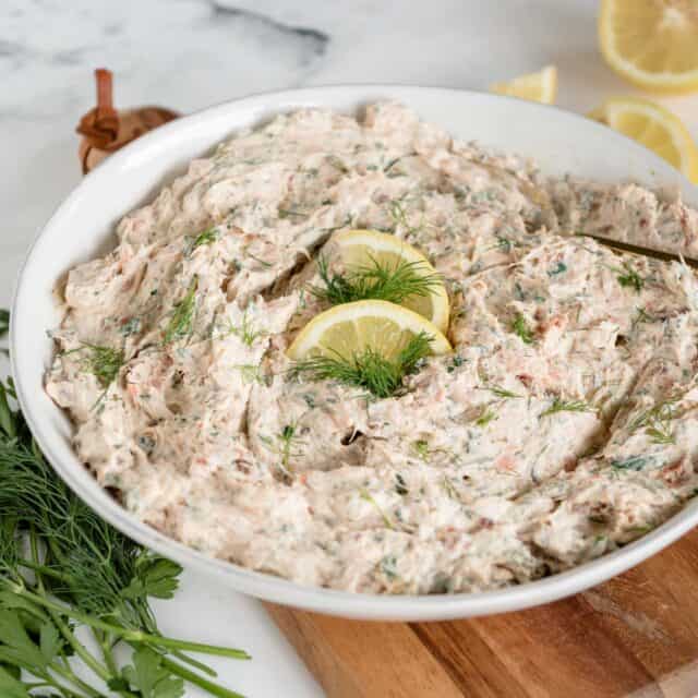 bowl of salmon dip from side view on cutting board on white background with a bunch of fresh herbs next to it