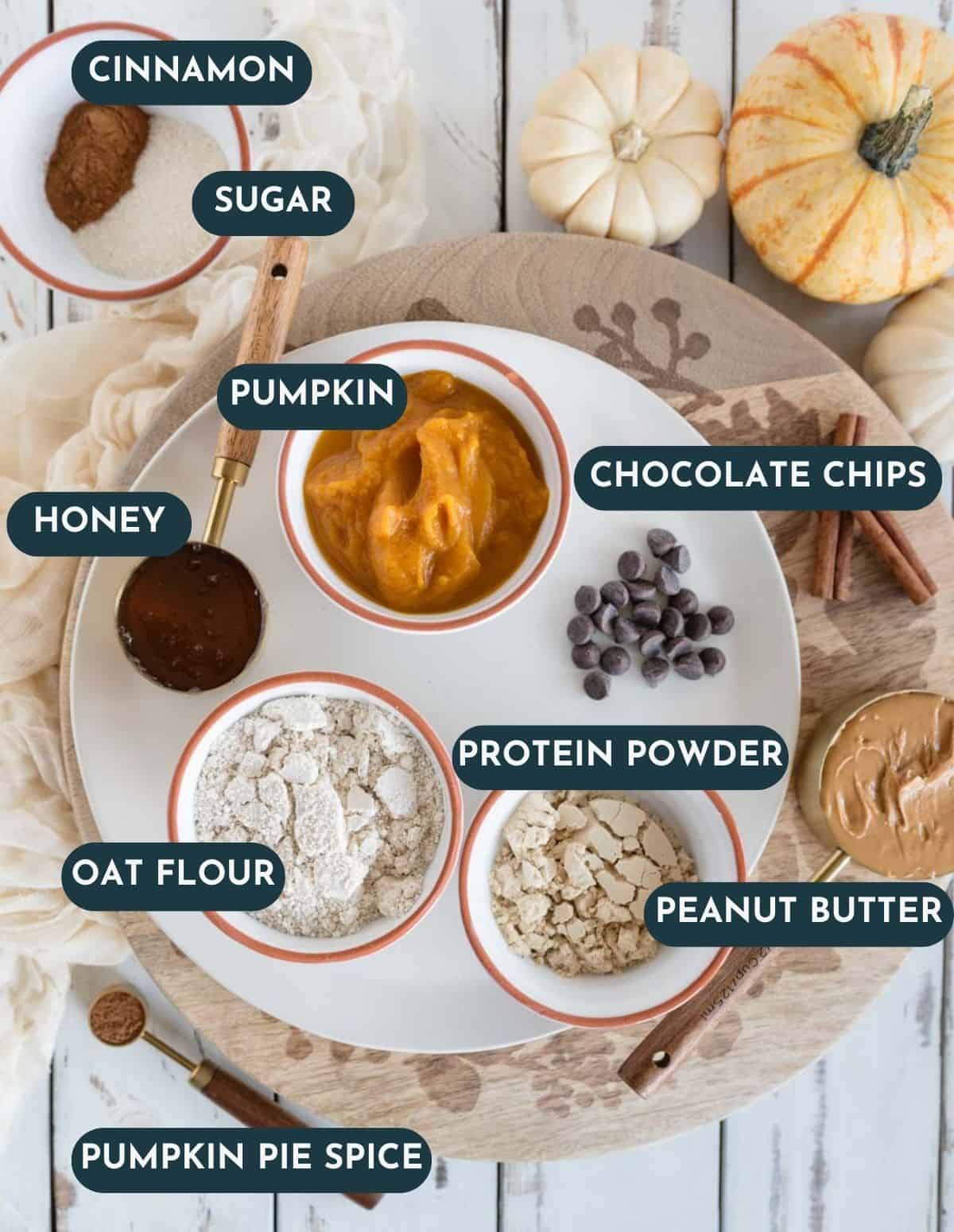 labeled ingredients in bowls and measuring cups cinnamon, sugar, honey, chocolate chips, oat flour, protein powder, pumpkin puree, and peanut butter.