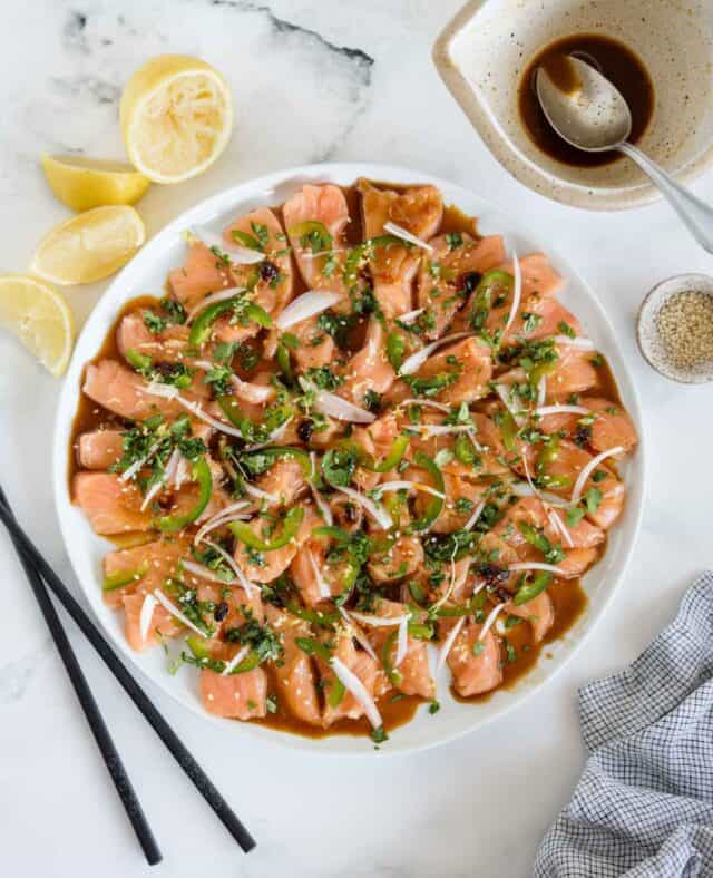 plate of sliced salmon topped with shallot, soy dressing, jalapeno and other toppings next to chopsticks, lemon and a bowl of sesame seeds