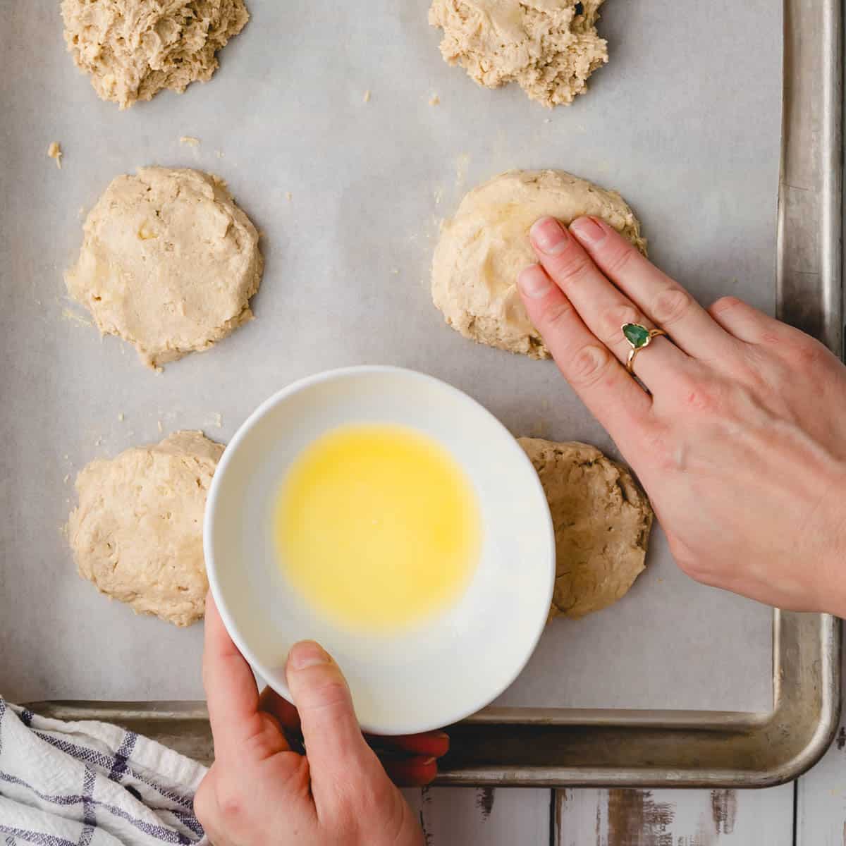 hand patting butter onto a biscuit on a baking sheet