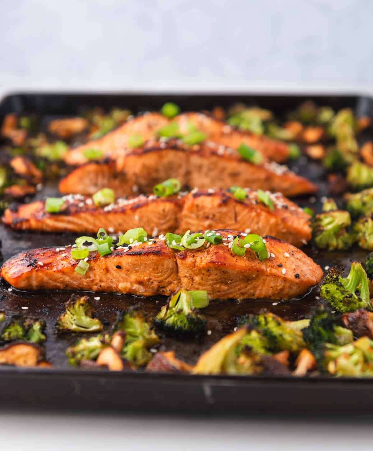 side view of baked salmon filets with broccoli and mushrooms with green onion and sesame seeds on top on a dark baking sheet