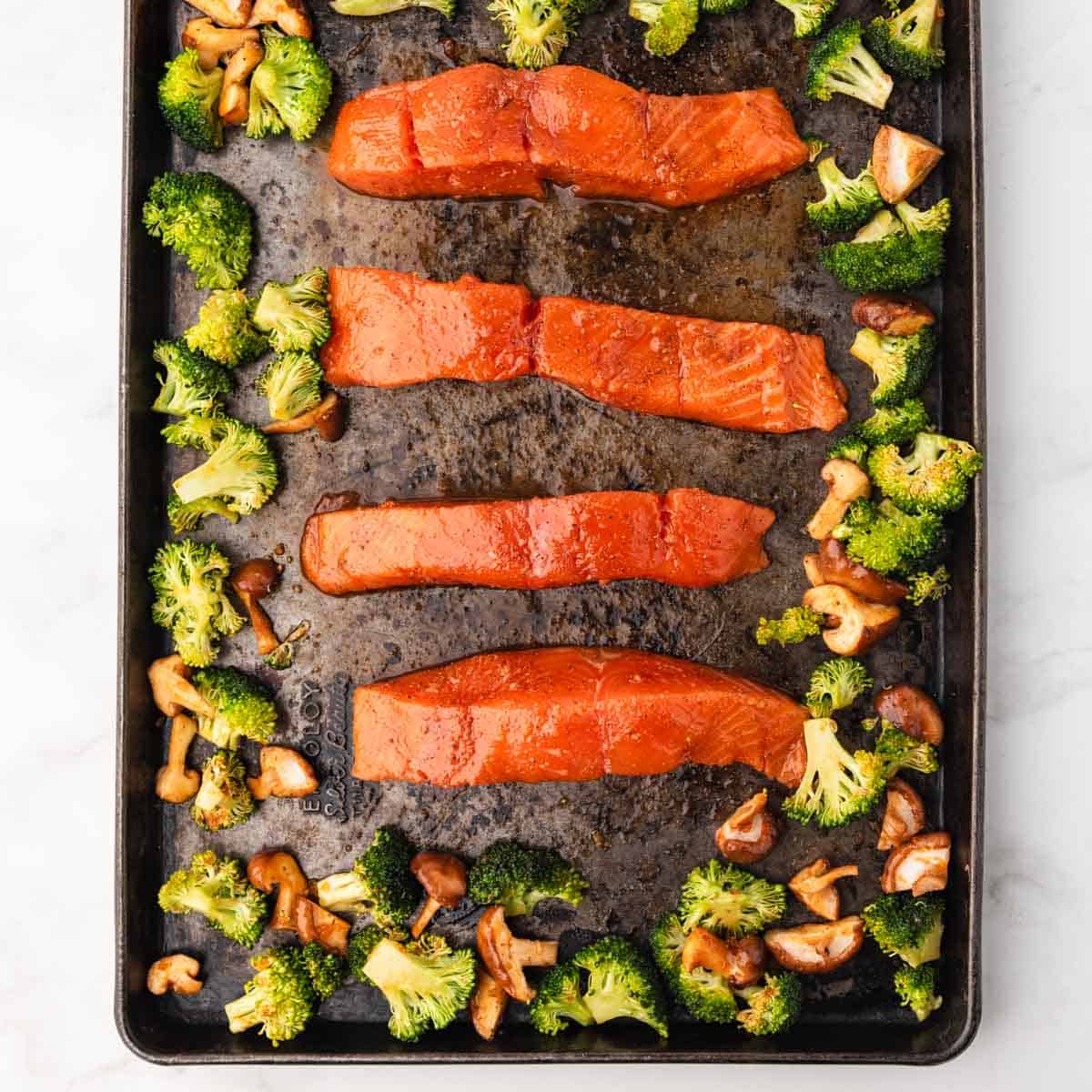 Salmon fillets on dark baking sheet surrounded by shiitake mushrooms and broccoli
