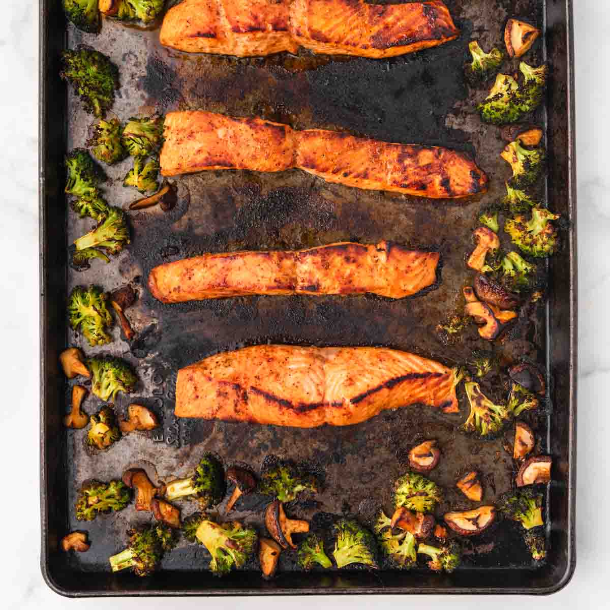 cooked salmon fillets on a dark baking sheet surrounded by shiitake mushrooms and broccoli