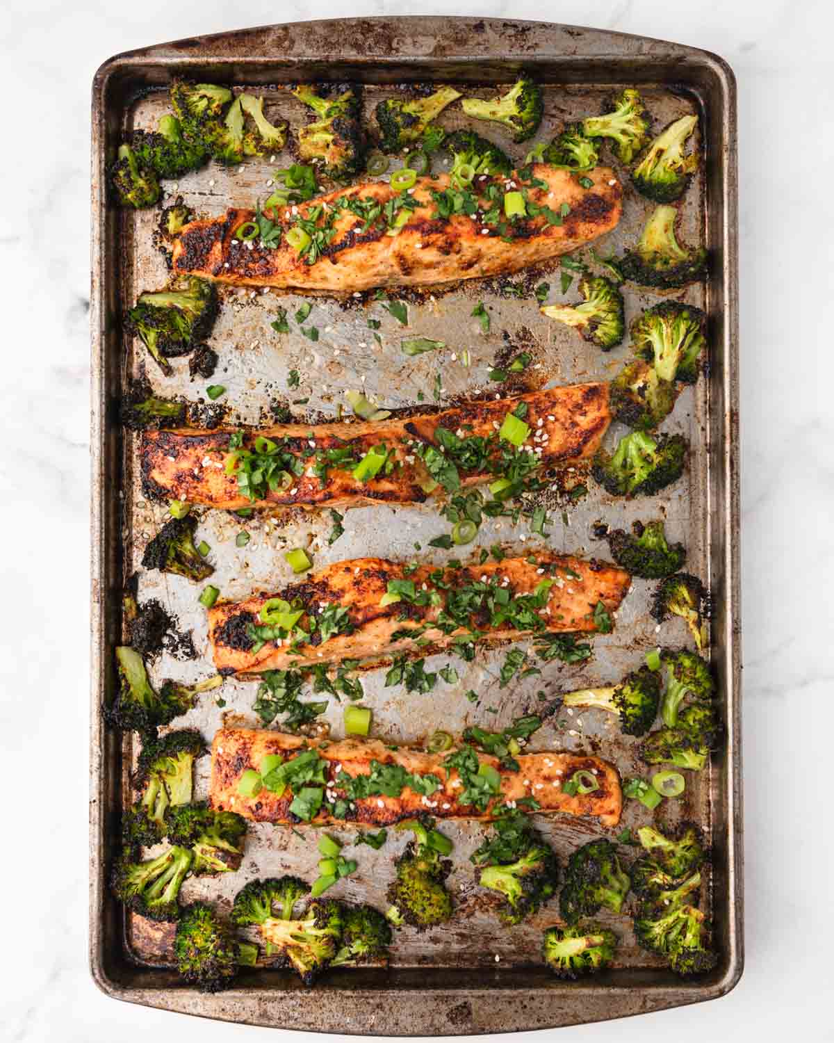 four salmon fillets laid out on a baking sheet topped with green onion, sesame seeds surrounded by broccoli