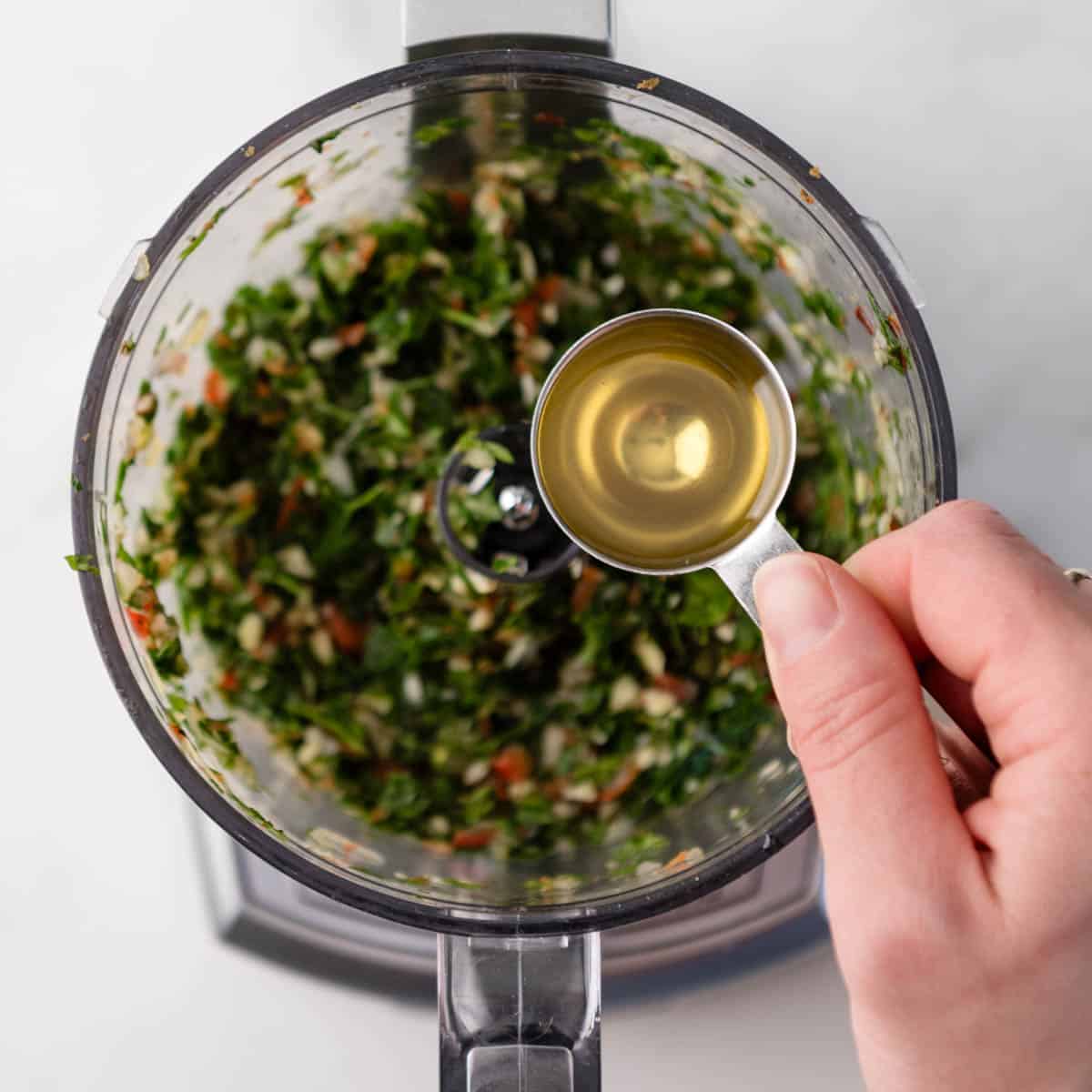Hand holding tablespoon of olive oil over food processor with blended herbs in it