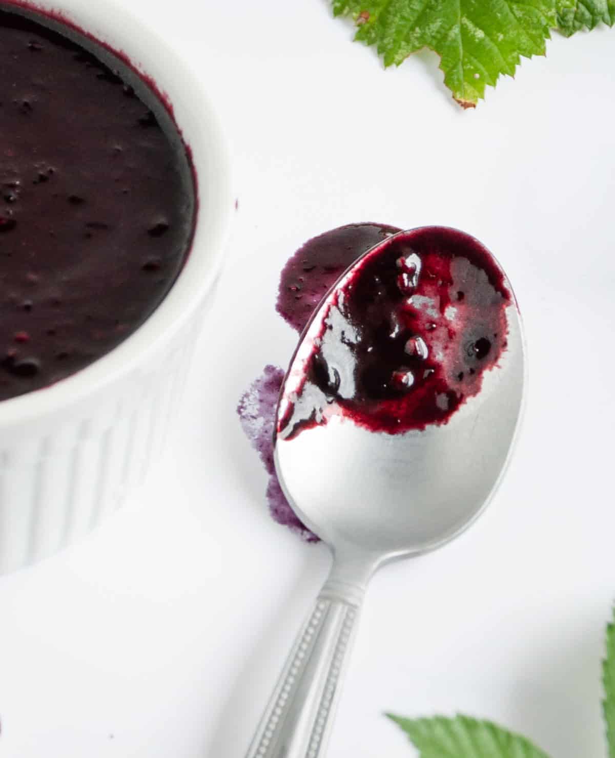 Close up of a spoon with blackberry salad dressing on it next to a larger container and leaves