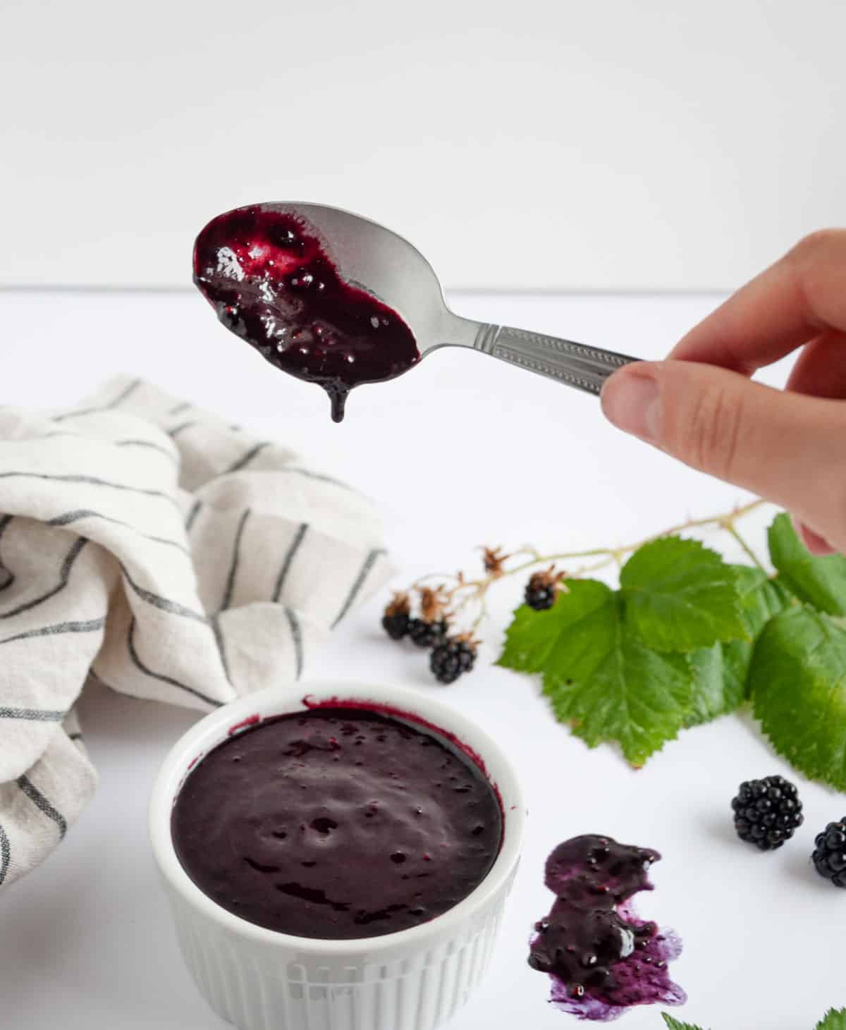 Hand holding spoon with dark sauce on it over a bowl with more in it and blackberries in background