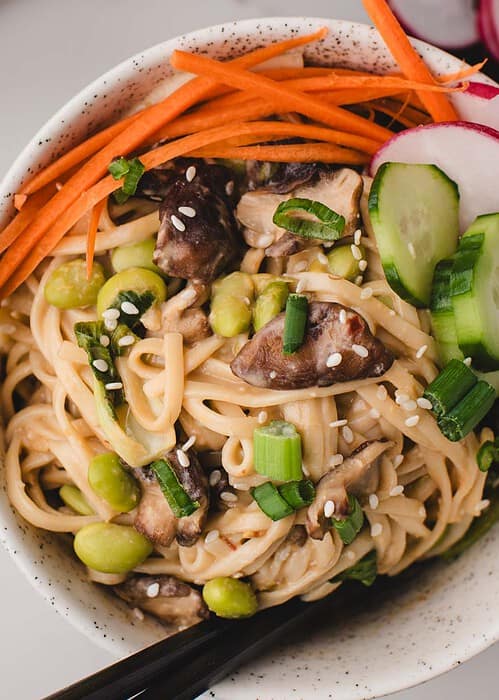close up bowl of miso noodles with chopsticks sticking out and carrot, edamame, sesame seeds and other veggies on top