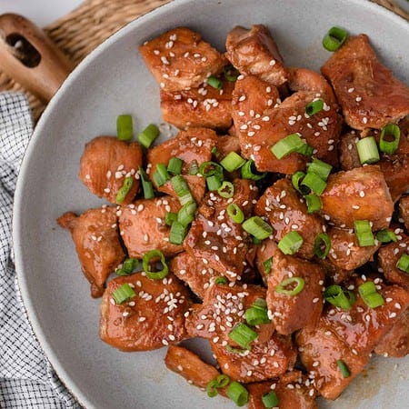 A plate of cooked chicken pieces with sesame seeds and chopped green onions on top, placed on a gray plate with a black and white napkin beside it.