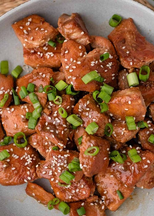 A plate of cooked chicken pieces with sesame seeds and chopped green onions on top, placed on a gray plate with a black and white napkin beside it.