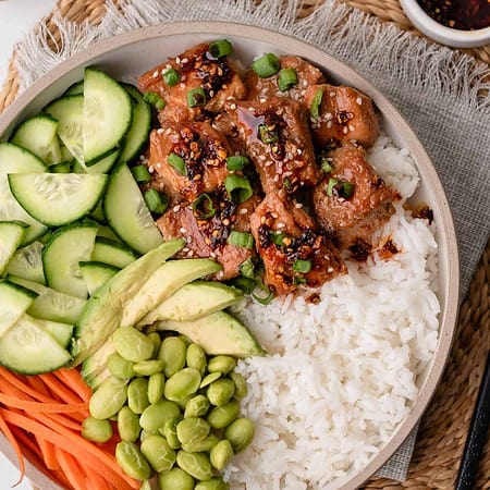 overhead of teriyaki salmon rice bowls with edamame, cucumber, avocado, and carrot on top of rice and saucy salmon pieces