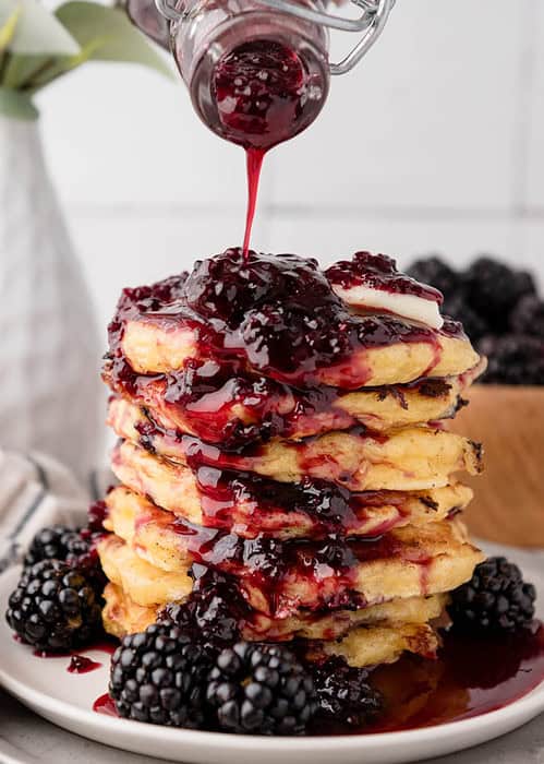 blackberry syrup being drizzled onto a stack of pancakes from the side with more fresh blackberries on the plate and background